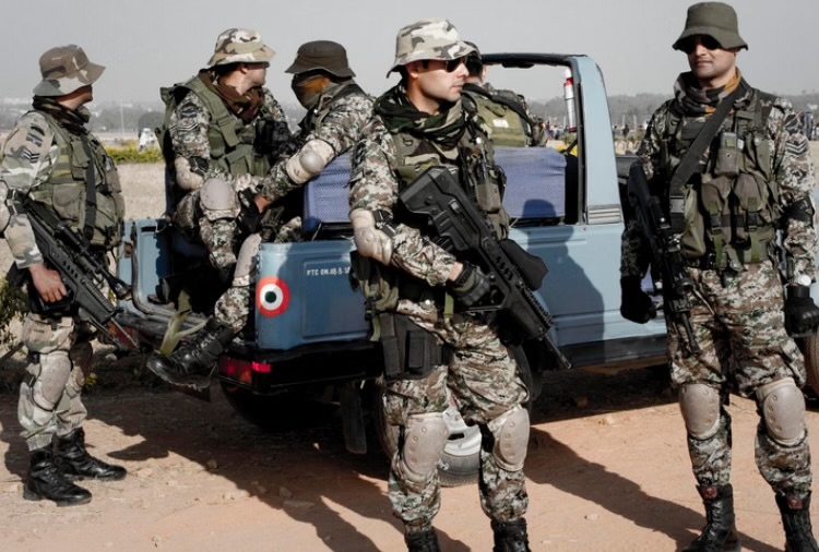 Commandos in camouflage, helmets and tactical vests with rifles standing guard beside a light-blue vehicle on a dusty airfield