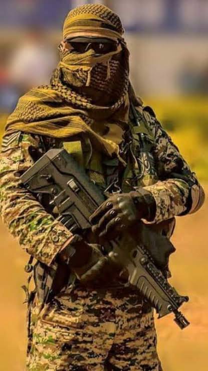 A masked commando wearing a shemagh, sunglasses and camouflage holding a rifle across the chest