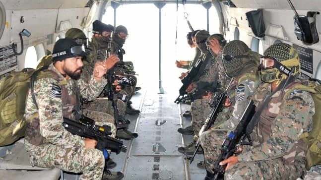 Commandos in camouflage, balaclavas and helmets seated inside a transport helicopter cabin with rifles